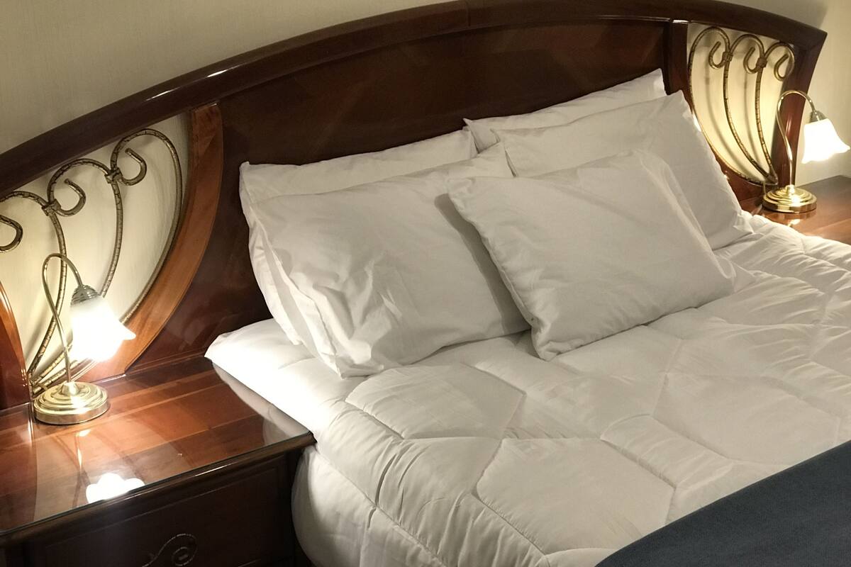 Ein ordentlich bezogenes Bett mit weißen Leinen und einem Holzkopfende mit geschwungenen Metalldekorationen. Auf jeder Seite des Bettes stehen je eine Nachttischlampe.