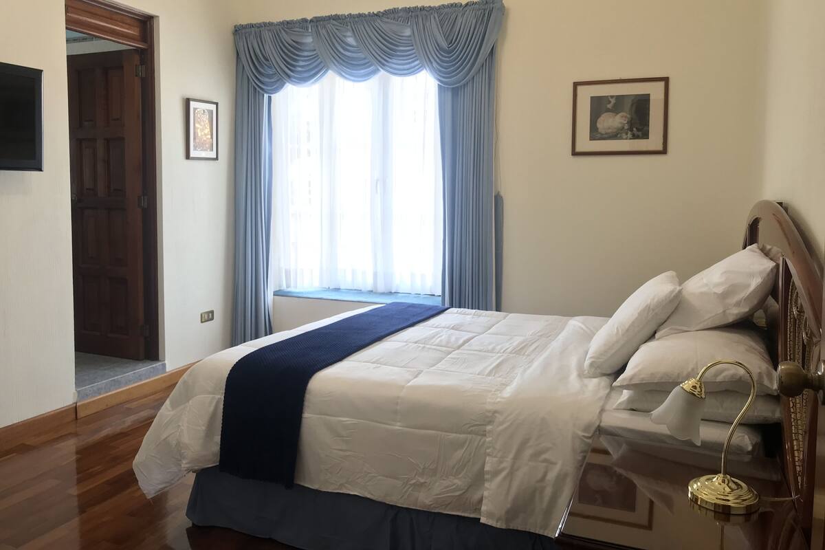 Ein ordentlich eingerichtetes Schlafzimmer mit Bett, Nachttischlampe, einem Fenster mit Vorhängen, einem gerahmten Bild an der Wand und einer Holztür.