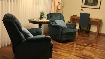 Um quarto com duas poltronas azul-escuras, uma pequena mesa redonda com uma planta, uma mesinha de cabeceira de madeira e um quadro pendurado na parede.