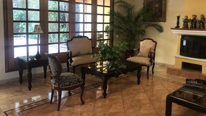 Een woonkamer met een open haard, twee fauteuils, een salontafel en een groot raam met uitzicht op groen.