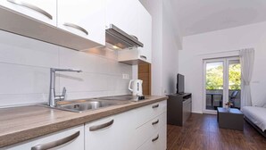 A modern kitchen with white cabinets, a stainless steel sink, and a built-in oven. There is a small seating area with a sofa and a coffee table. A television is mounted on the wall.
