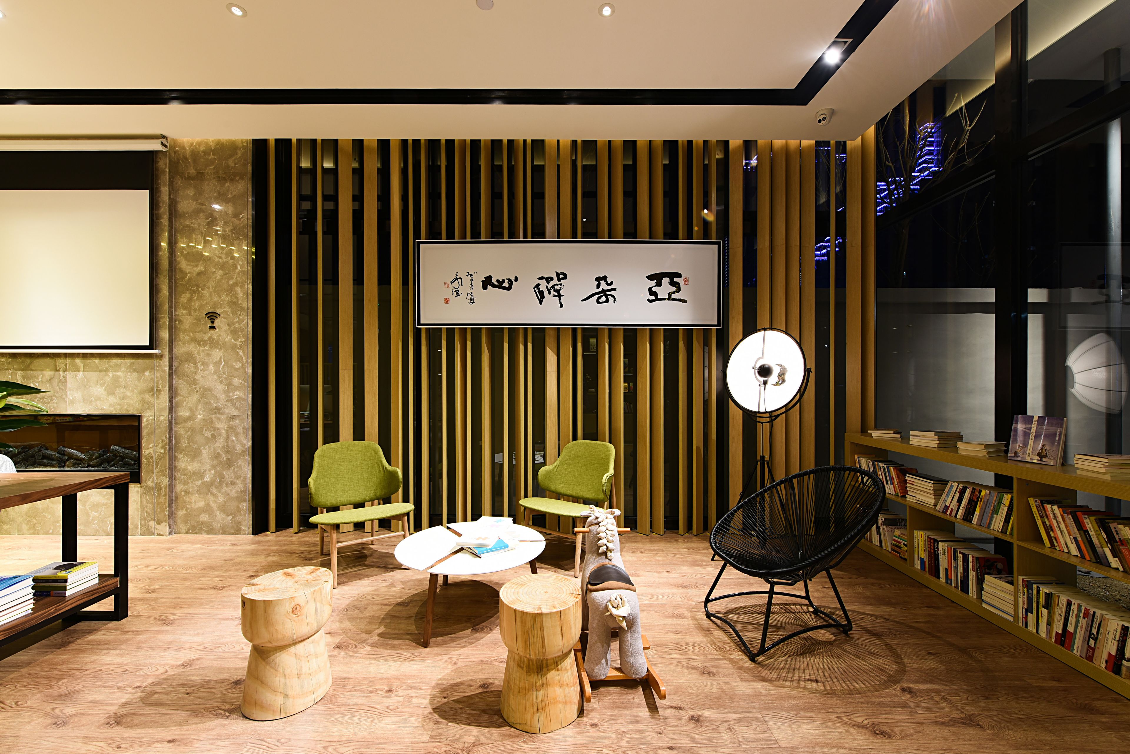 A modern interior with a wooden floor, green chairs, a round table, a black chair, and a bookshelf.