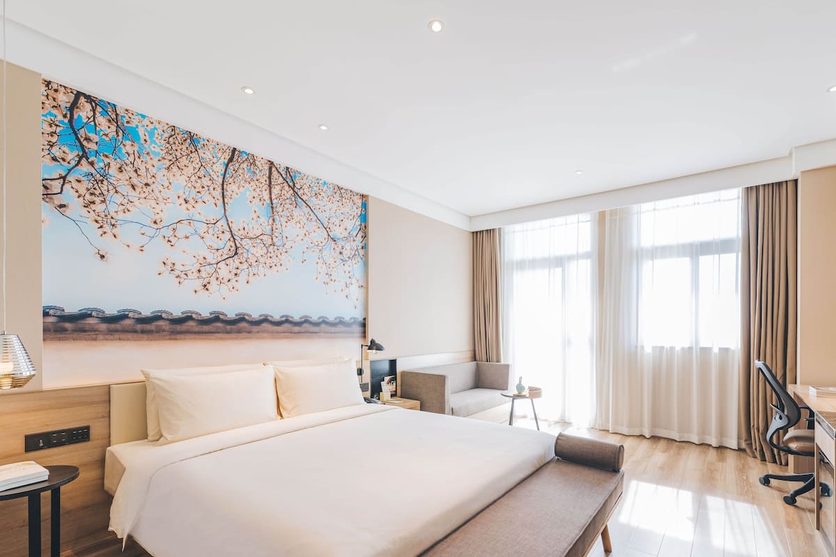 Ein modernes Hotelzimmer mit einem großen Bett, einem Schreibtisch und einem Stuhl. Es gibt ein großes Fenster mit Vorhängen, ein Bild an der Wand und eine Deckenleuchte.