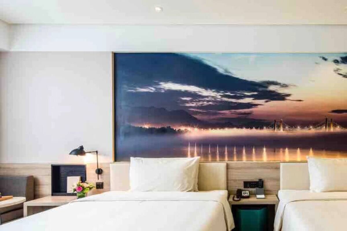 Ein Hotelzimmer mit zwei Betten, einem großen Gemälde mit Sonnenuntergang über Wasser und einem hölzernen Kopfende.