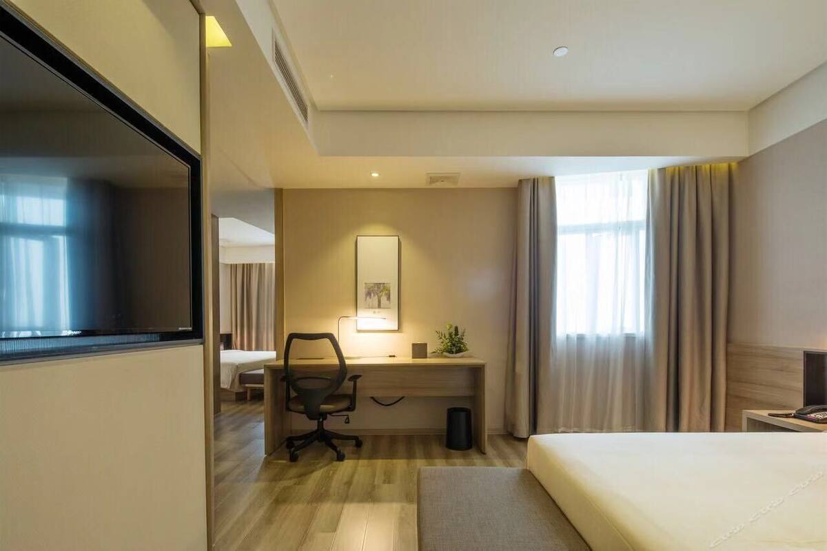 Ein Hotelzimmer mit Bett, Schreibtisch mit Stuhl, einem kleinen Tisch, einer Nachttischlampe, einem Spiegel und einem Fenster mit Vorhängen.