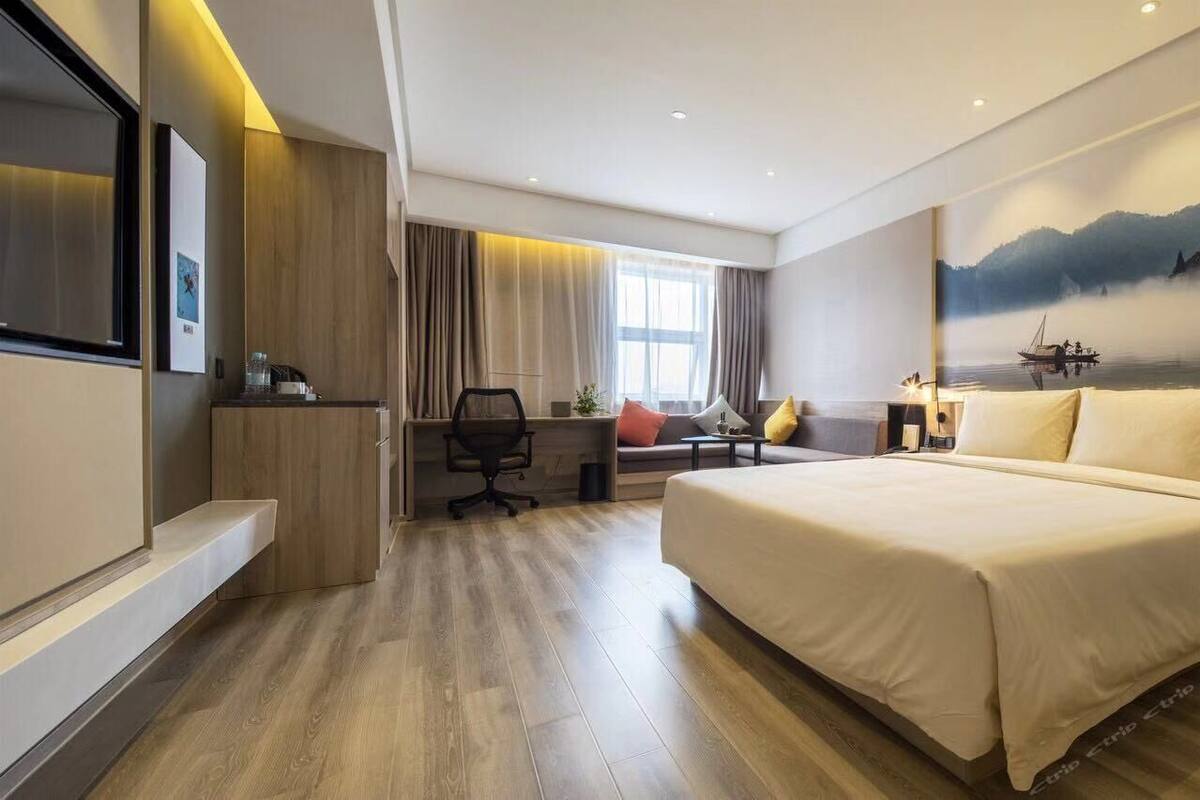 Ein modernes Hotelzimmer mit einem großen Bett, einem Schreibtisch mit Stuhl, einer Couch und einem großen Fenster mit Vorhängen.