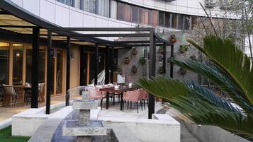 A modern building with a curved facade, outdoor seating area with tables and chairs, a water feature, and potted plants.