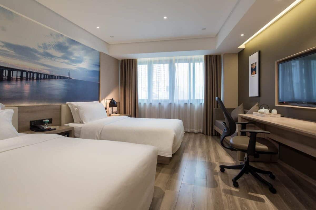 Habitación de hotel con dos camas, un escritorio, una silla, un televisor y un ventanal grande con cortinas.