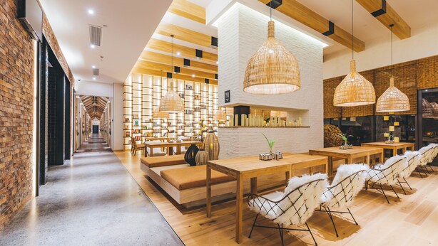 Un interior moderno con un largo pasillo que conduce a una zona de estar con mesas y sillas, vigas de madera en el techo y una pared de ladrillo a un lado.