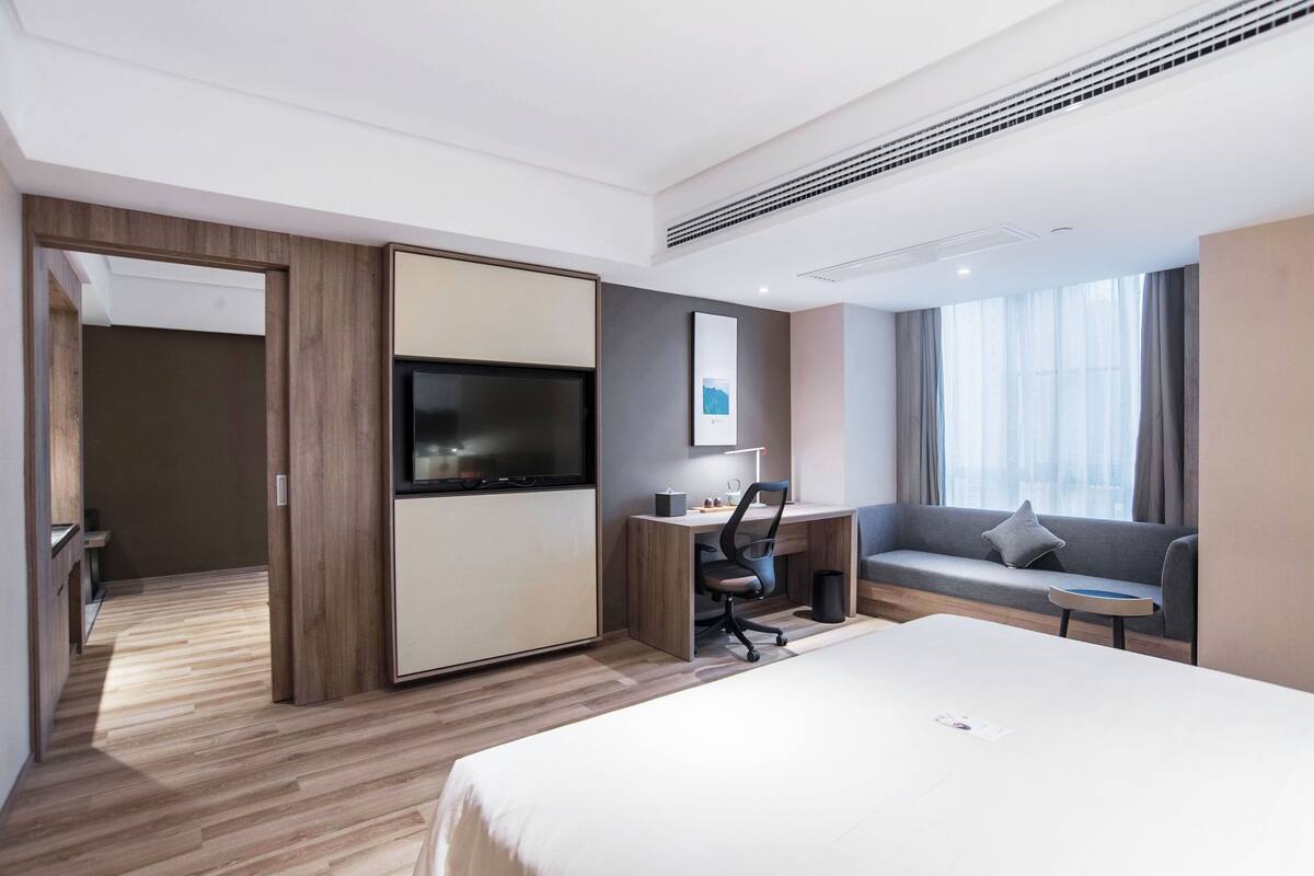 A hotel room with a bed, a desk with a chair, a TV, a sofa, and a window with curtains.