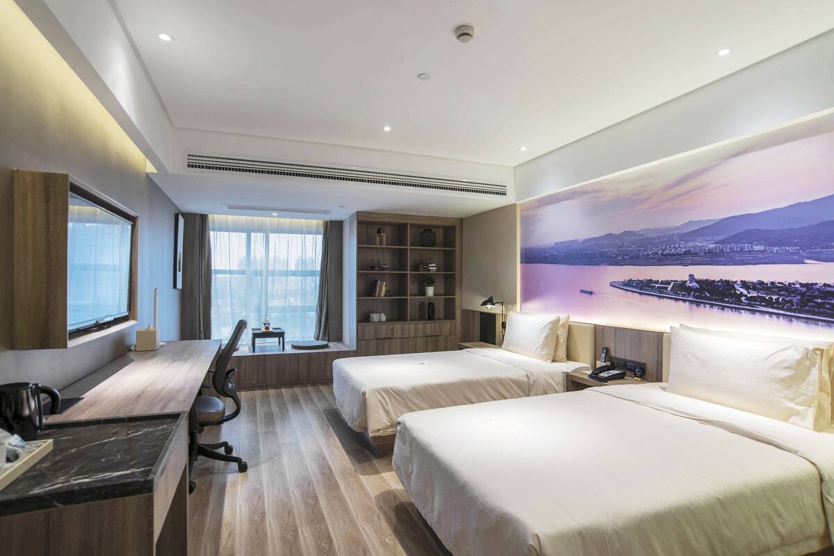 A hotel room with two beds, a desk, a bookshelf, and a large window.