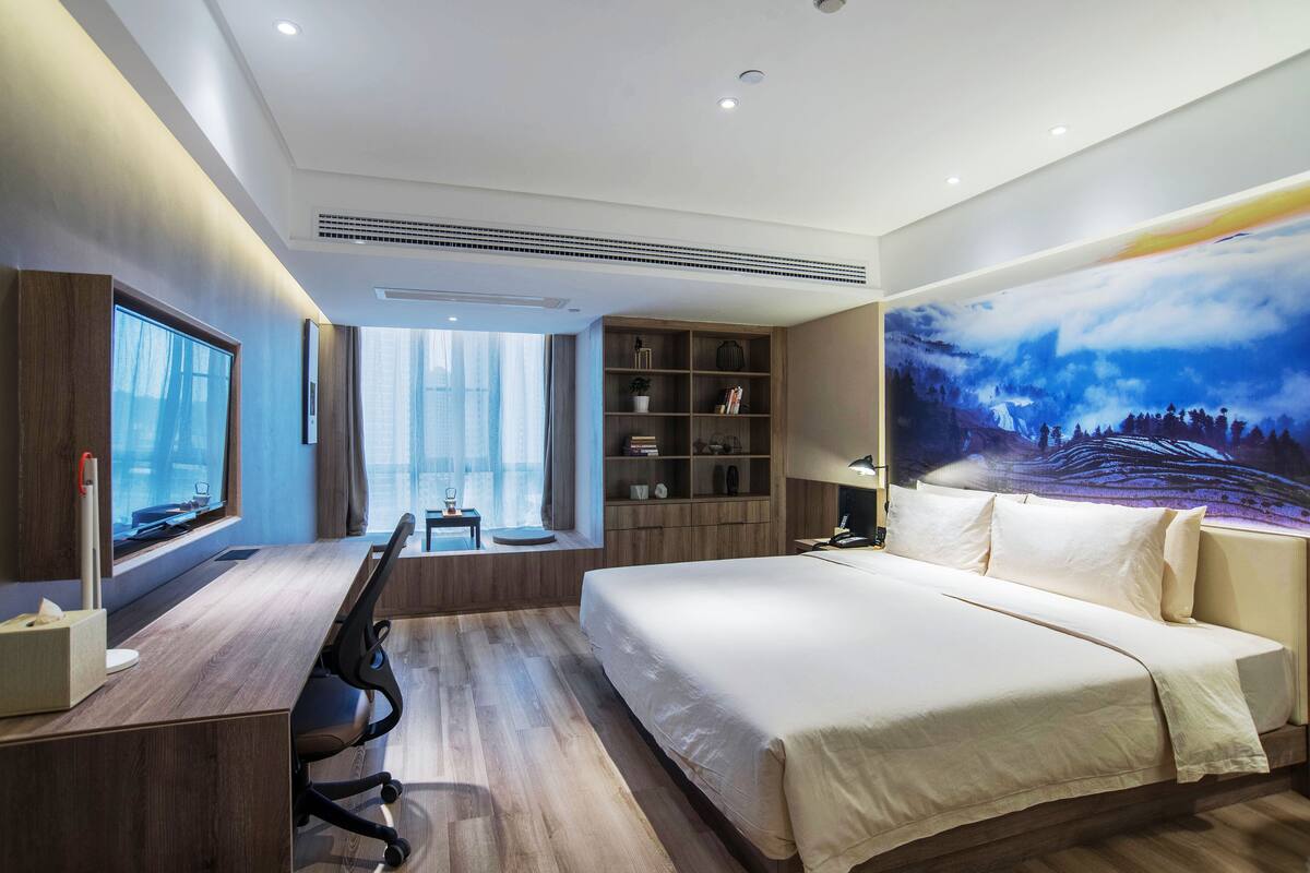 A modern hotel room with a large bed, a desk with a computer, a bookshelf, and a large window with curtains.