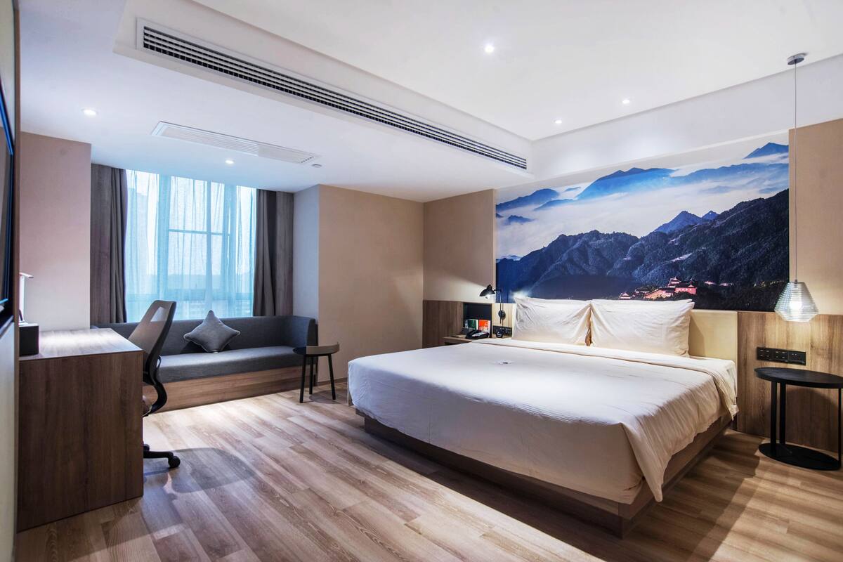 A modern hotel room with a large bed, a desk, a chair, a sofa, and a large window.