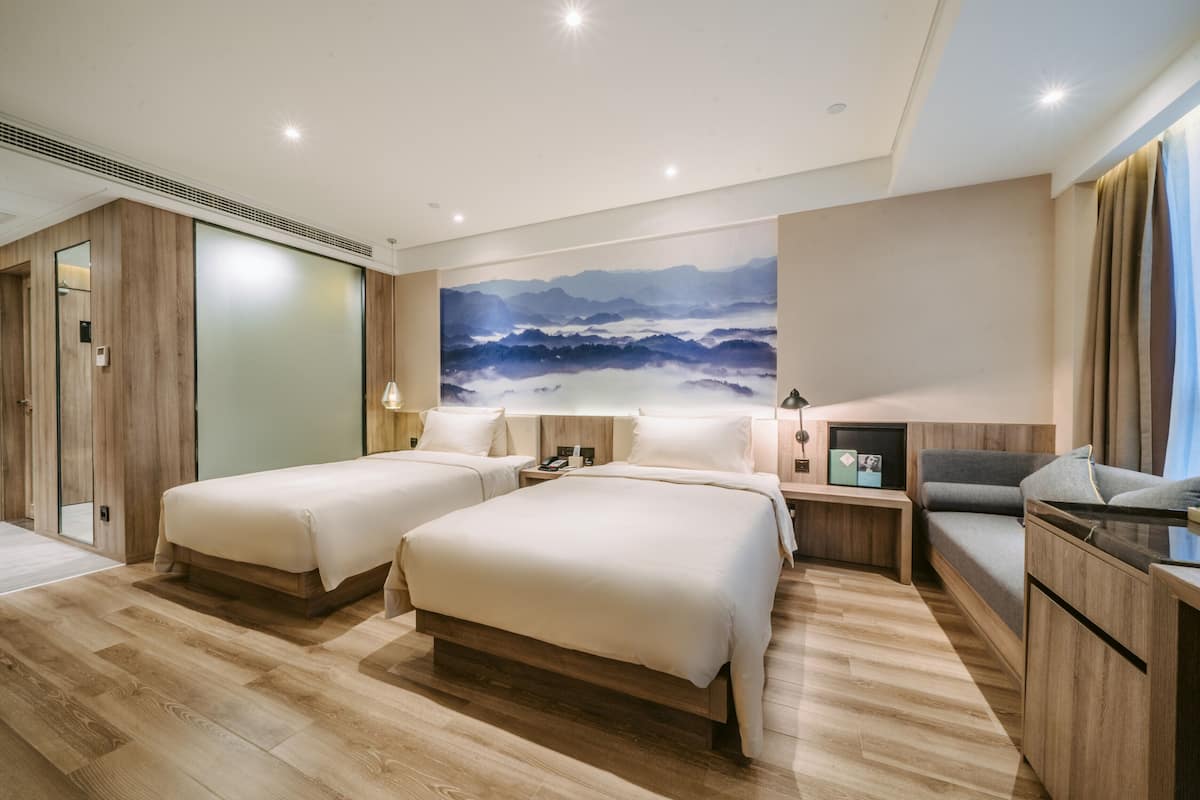 A hotel room with two beds, a desk, a chair, and a large painting of mountains.