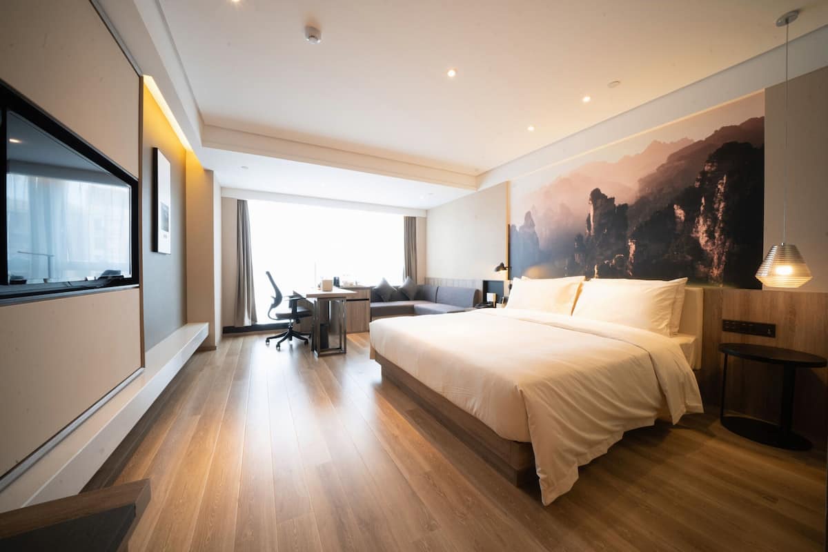 A modern hotel room with a large bed, a desk, a chair, a TV, and a painting of a mountain landscape.