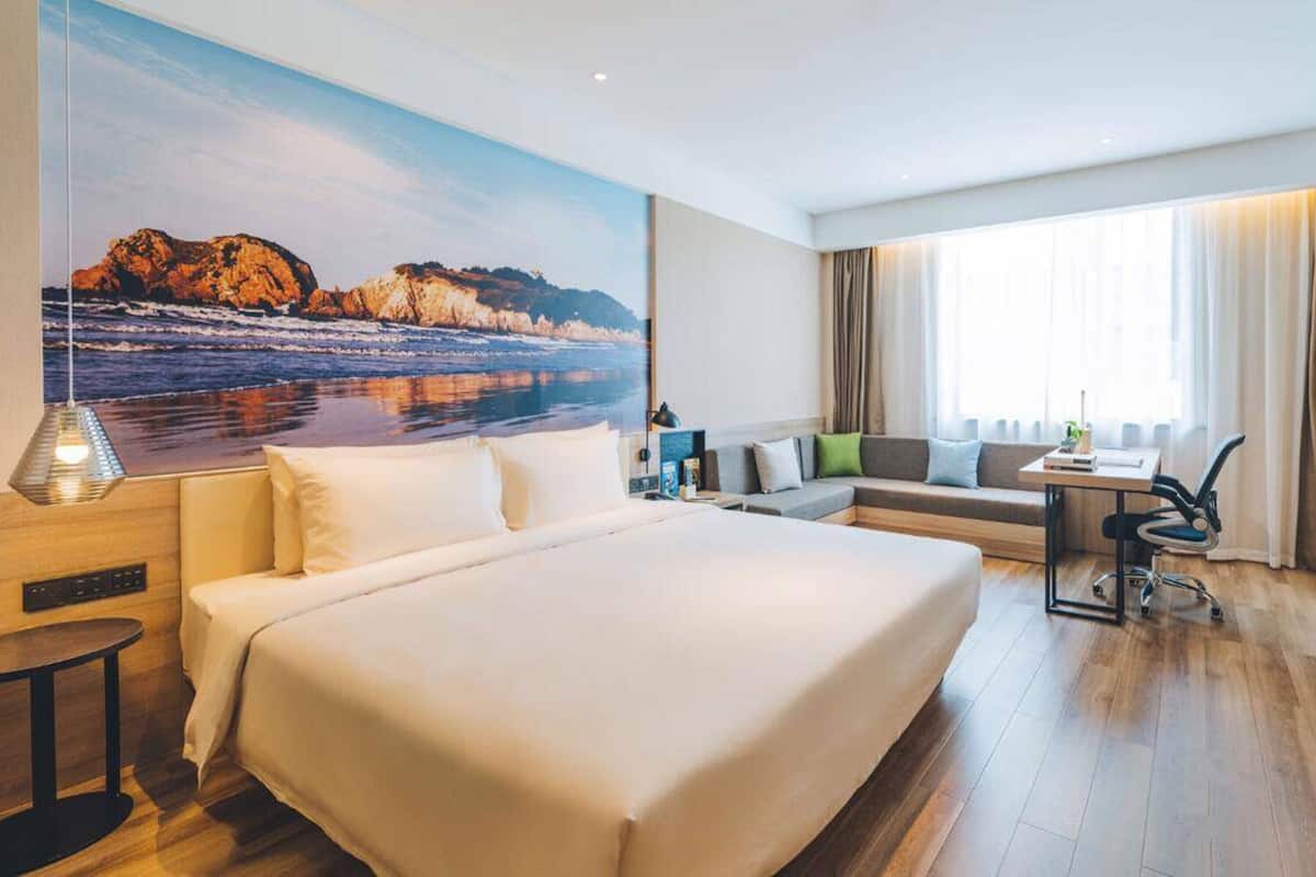 A hotel room with a large bed, a sofa, a desk, and a painting of a coastal scene.