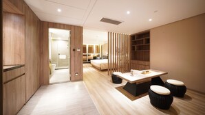 Une chambre d’hôtel moderne avec un plancher en bois, un lit, un bureau avec des tabourets et une salle de bain visible à travers une porte ouverte.