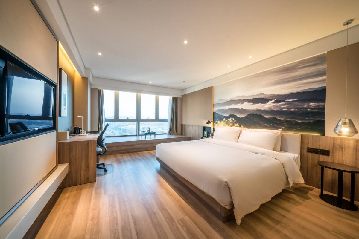 A modern hotel room with a large bed, a desk, a chair, a TV, and a large window with a view.