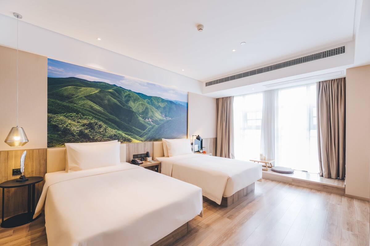 Habitación de hotel con dos camas, un ventanal con cortinas, un cuadro colgado en la pared de un paisaje y una mesita con una lámpara.