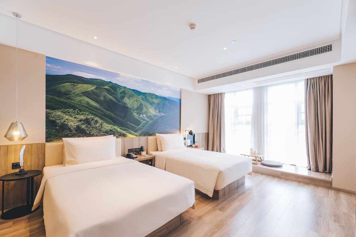 Une chambre d’hôtel avec deux lits, une grande fenêtre avec des rideaux, un tableau représentant un paysage accroché au mur, et une petite table avec une lampe.