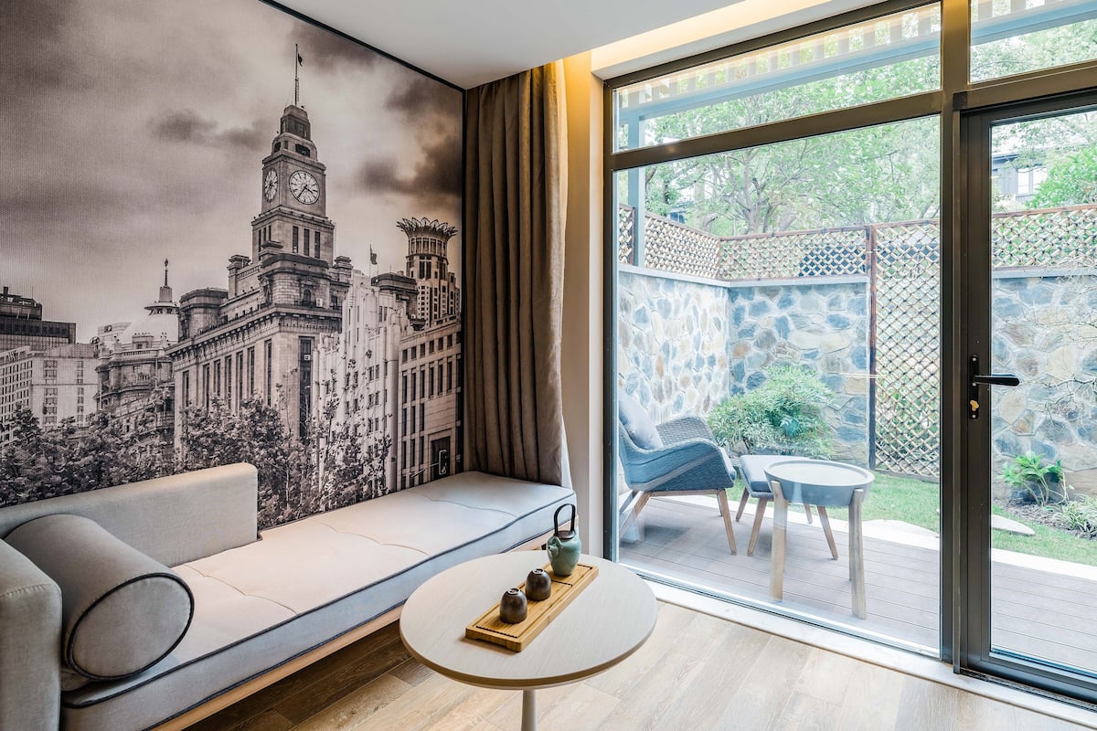 Ein modernes Wohnzimmer mit einer Couch, einem kleinen runden Tisch und einem großen Fenster mit Blick auf einen Garten sowie einem Wandgemälde mit Stadtansicht.