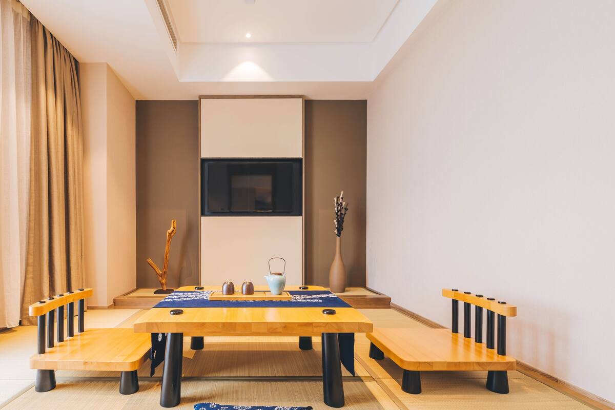 A modern dining area with a wooden table and benches, a flat-screen TV mounted on the wall, and a vase with dried branches on a side table.