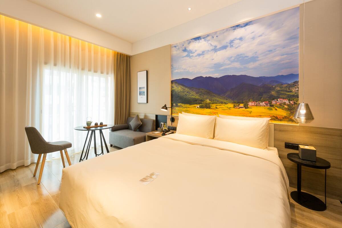 A hotel room with a large bed, a desk with a chair, a small table, and a painting of a landscape.