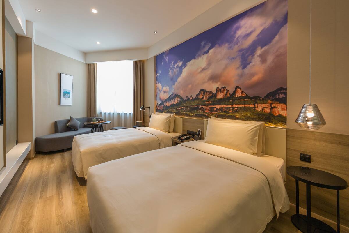 A hotel room with two beds, a desk, a chair, a TV, and a large mountain landscape painting on the wall.