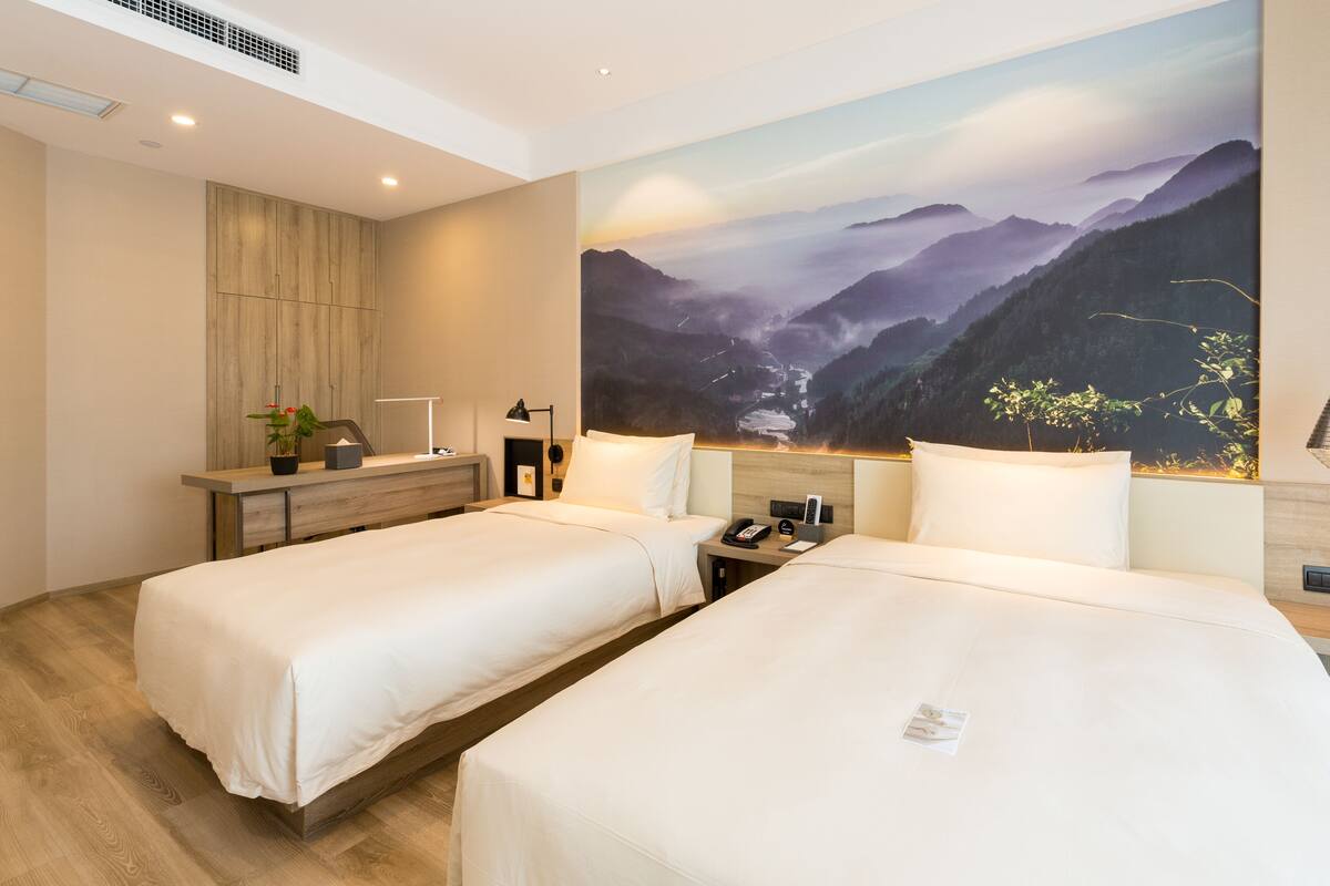 A hotel room with two beds, a desk, a nightstand, a painting of a mountain landscape, and a hanging pendant light.