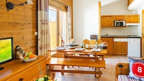 A cozy room with a wooden table, benches, a bed, a TV, and a kitchen area with wooden cabinets and appliances. - Appartement sympa et équipé, proche de le Navette (Samoëns)