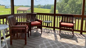 A covered outdoor area with wooden chairs and a small table, overlooking a green landscape.