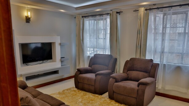 A living room with a flat-screen TV, two brown sofas, a beige rug, and a window with sheer curtains. - Esterina Suites (Nairobi)