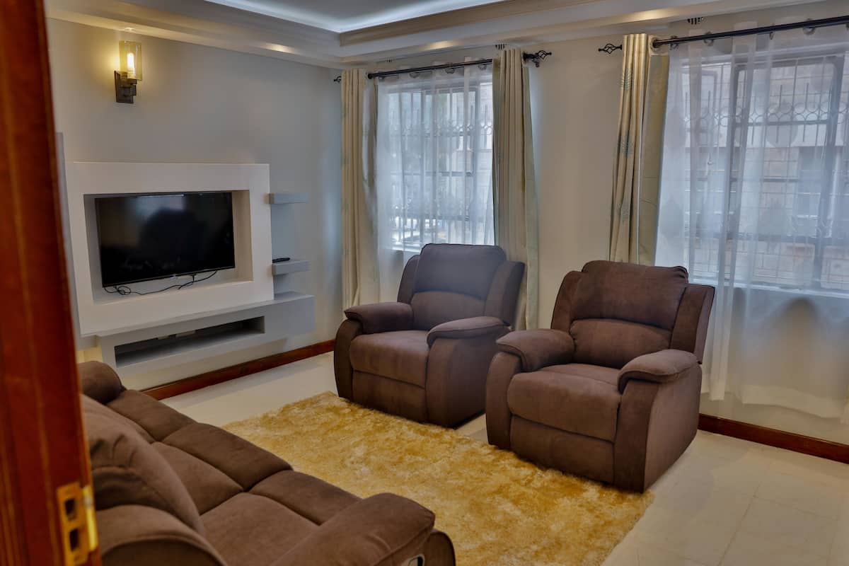 A living room with a flat-screen TV, two brown sofas, a beige rug, and a window with sheer curtains.