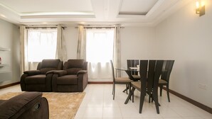 A living room with a brown sofa, a dining table with chairs, and a large window with curtains. - Esterina Suites (Nairobi)