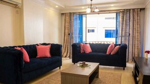 Ein Wohnzimmer mit zwei dunkelblauen Sofas, einem Holztisch und Blick auf ein Gebäude durch offene Vorhänge.