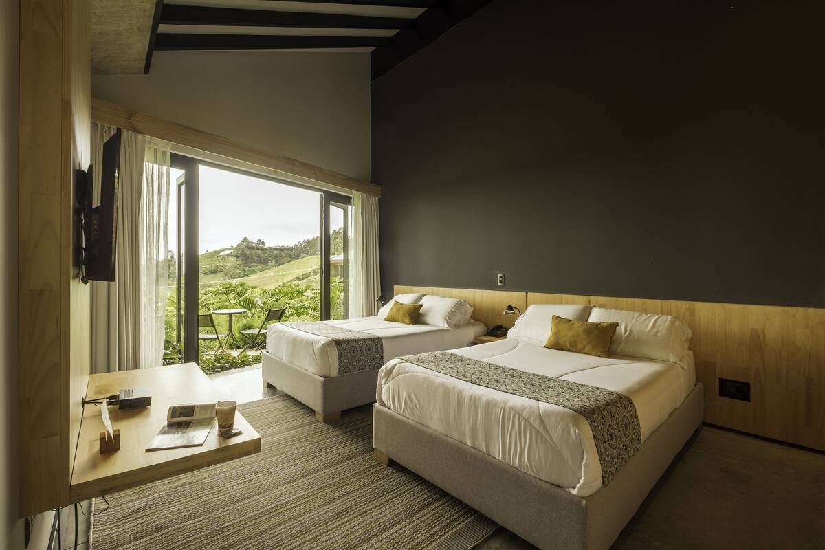 A hotel room with two beds, a desk, and a large window with a view of greenery.