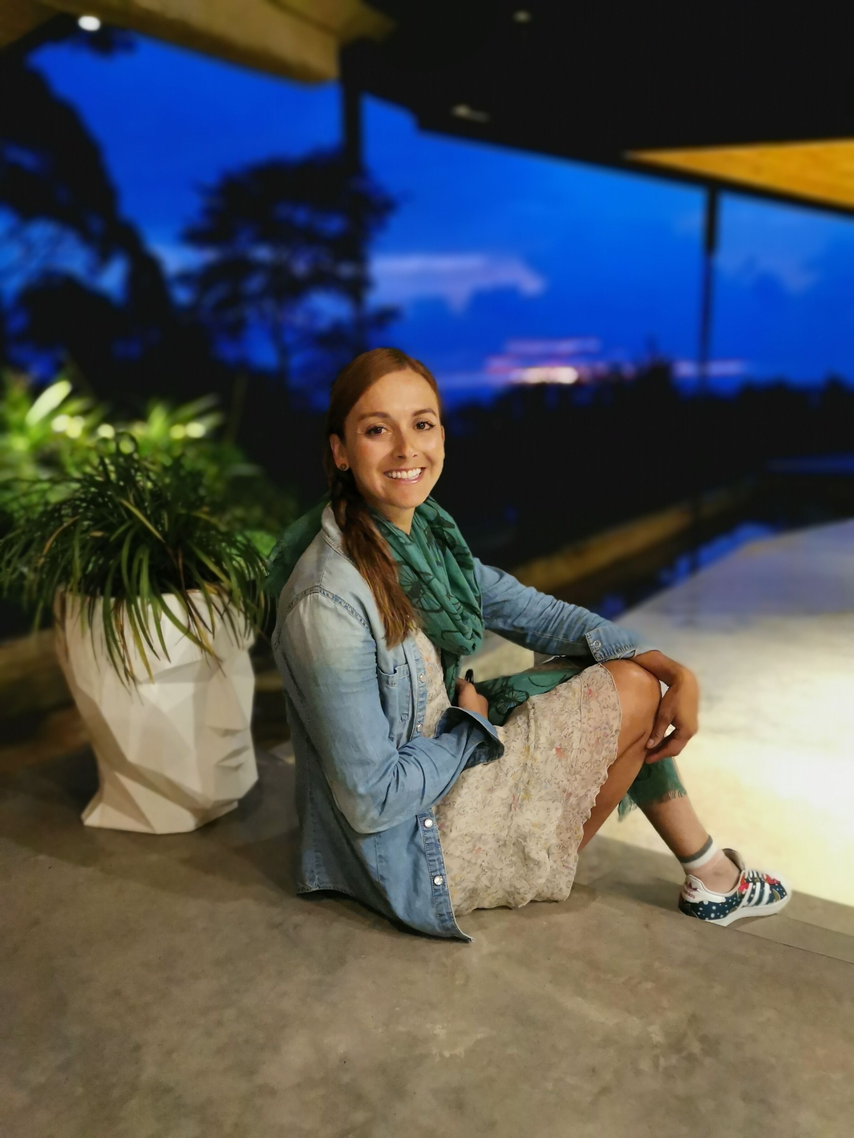 Una persona sentada en el suelo junto a una planta en maceta, en un espacio interior con vista al exterior.