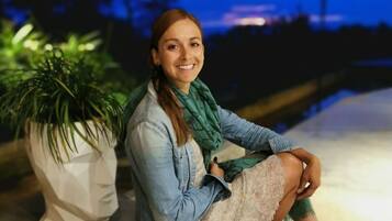 Una persona sentada en el suelo junto a una planta en maceta, en un espacio interior con vista al exterior.