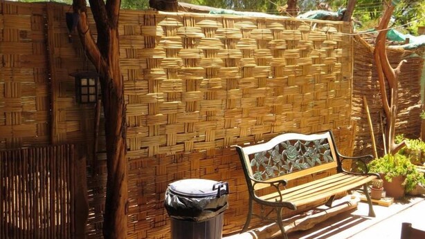 Pared de mimbre, un banco y un cubo de basura en un espacio exterior soleado.