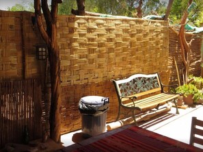 Pared de mimbre, un banco y un cubo de basura en un espacio exterior soleado.