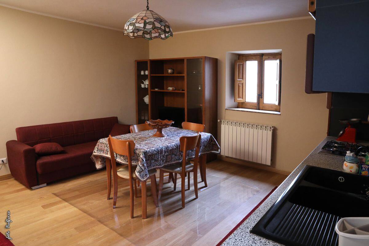 A compact living space with a dining table, chairs, a red sofa, a cabinet, a window, and a kitchenette.