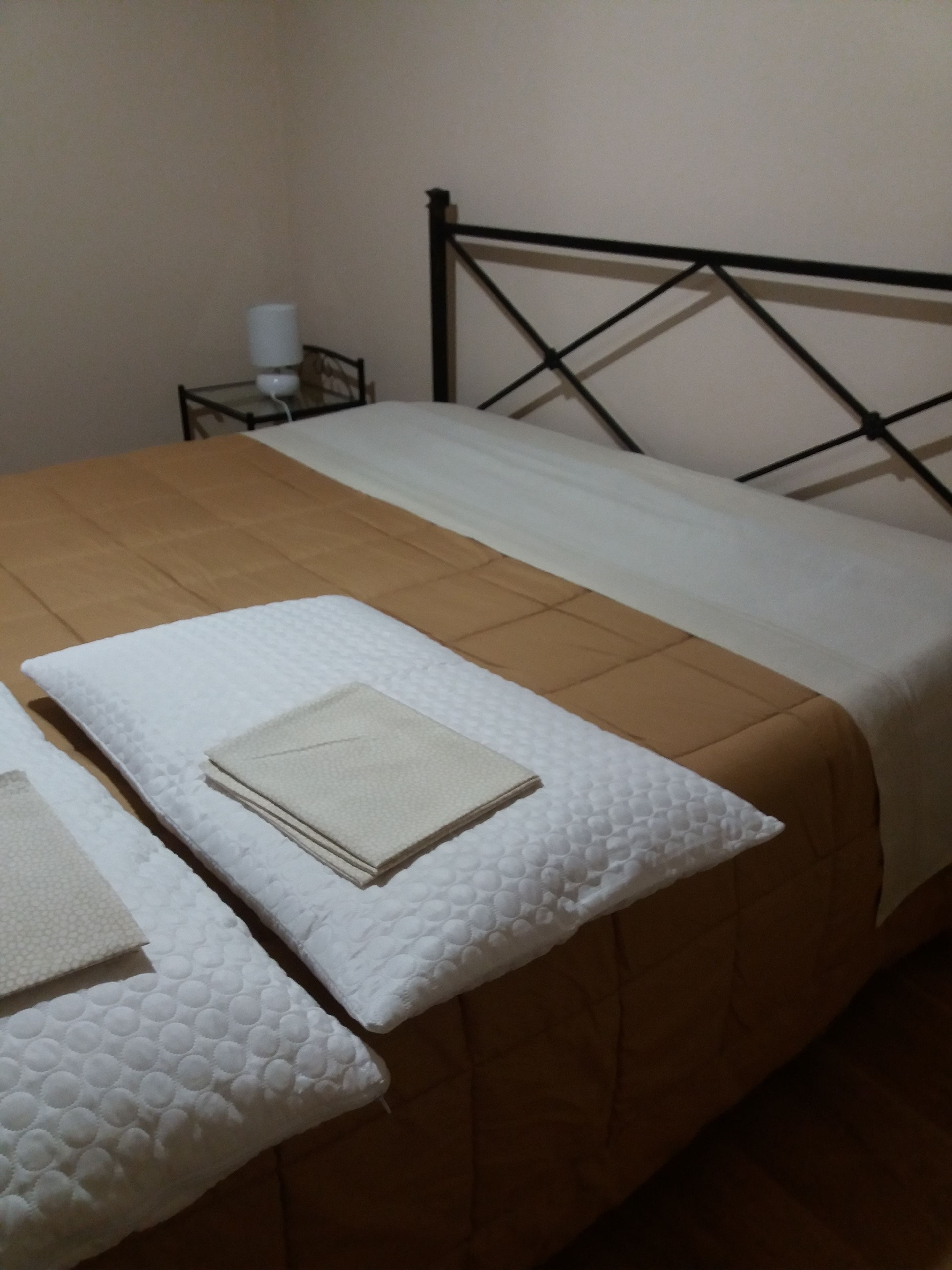 Een netjes opgemaakt bed met een bruine sprei en witte kussens, een klein nachtkastje met een lamp, en een eenvoudige muur.