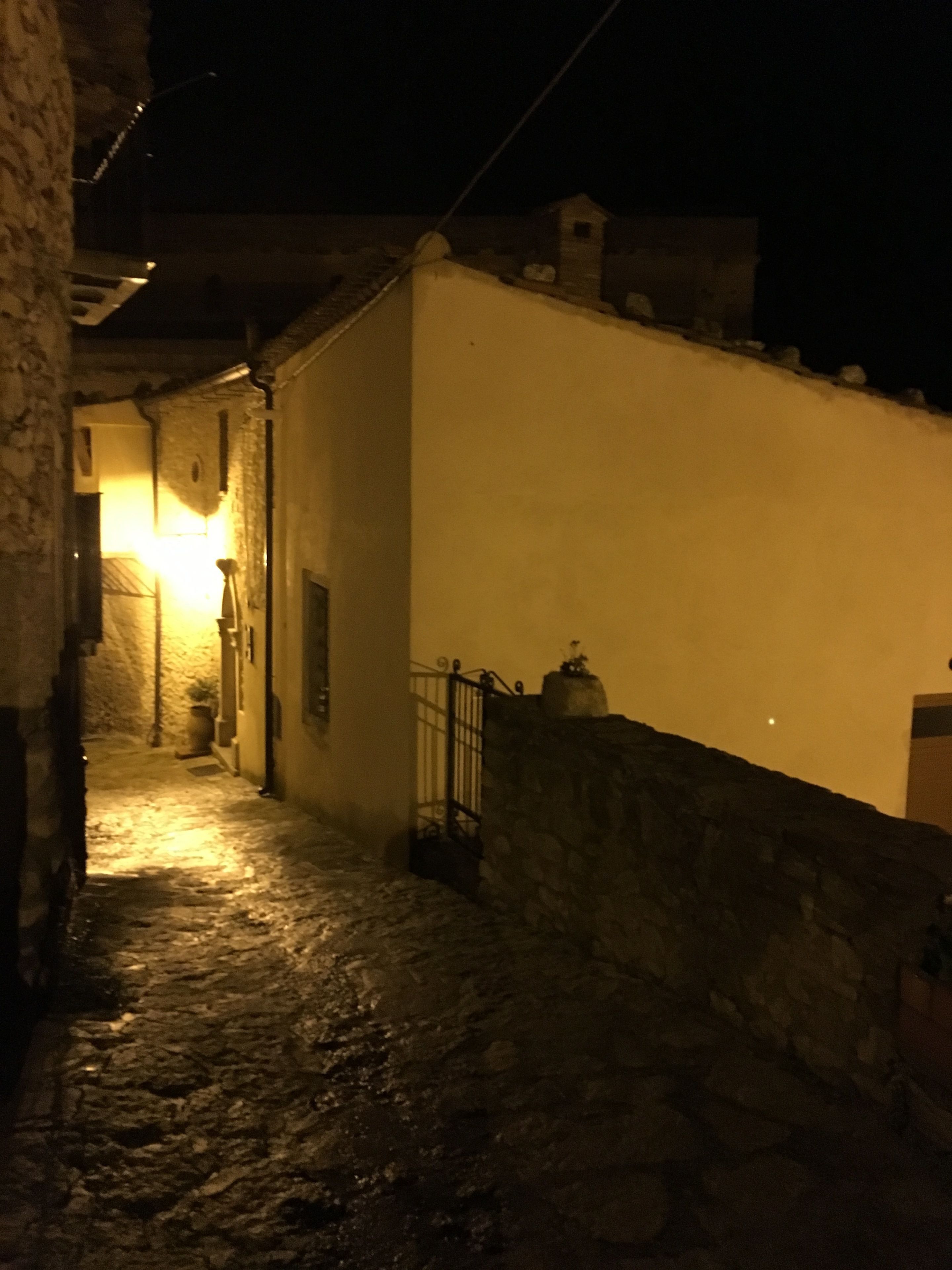 Een smalle geplaveide straat 's nachts, met een wit gebouw aan één kant en een stenen muur aan de andere kant.