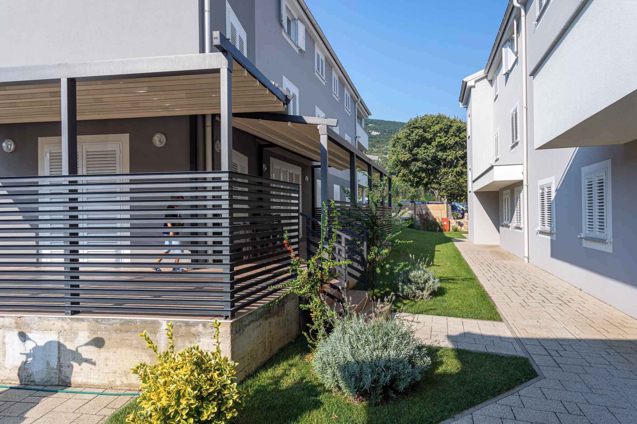 Un edificio residencial con un área exterior techada, un sendero pavimentado y un pequeño jardín con arbustos y césped.