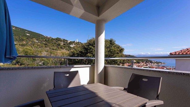Un balcon avec une table et des chaises, donnant sur une ville côtière et des collines.