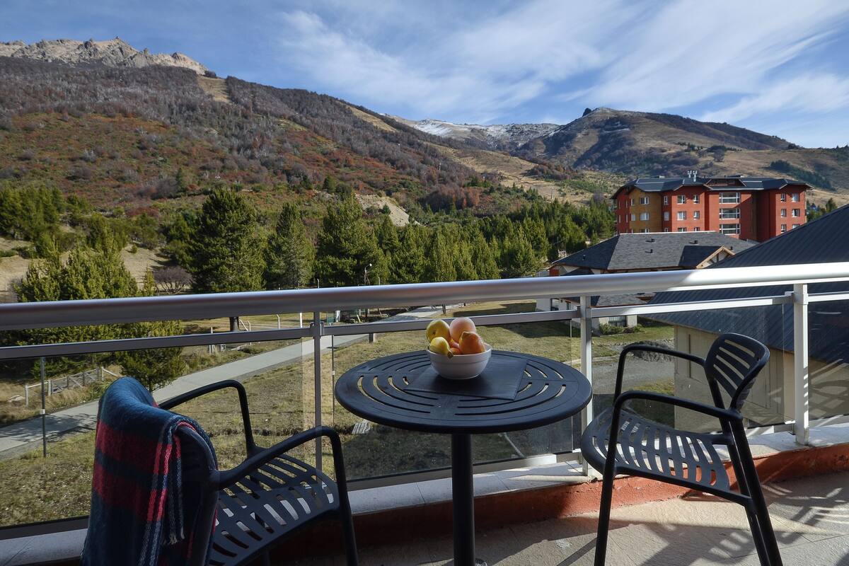 Ein Balkon mit Tisch und Stühlen, der auf eine gebirgige Landschaft und ein Gebäude blickt.