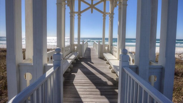 Un paseo marítimo de madera que lleva a un quiosco blanco con columnas, con vista a una playa.
