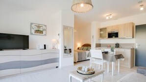 Ett modernt vardagsrum med en platt-TV, en soffa, ett matbord med stolar, en köksdel med träskåp och en hängande lampa.
