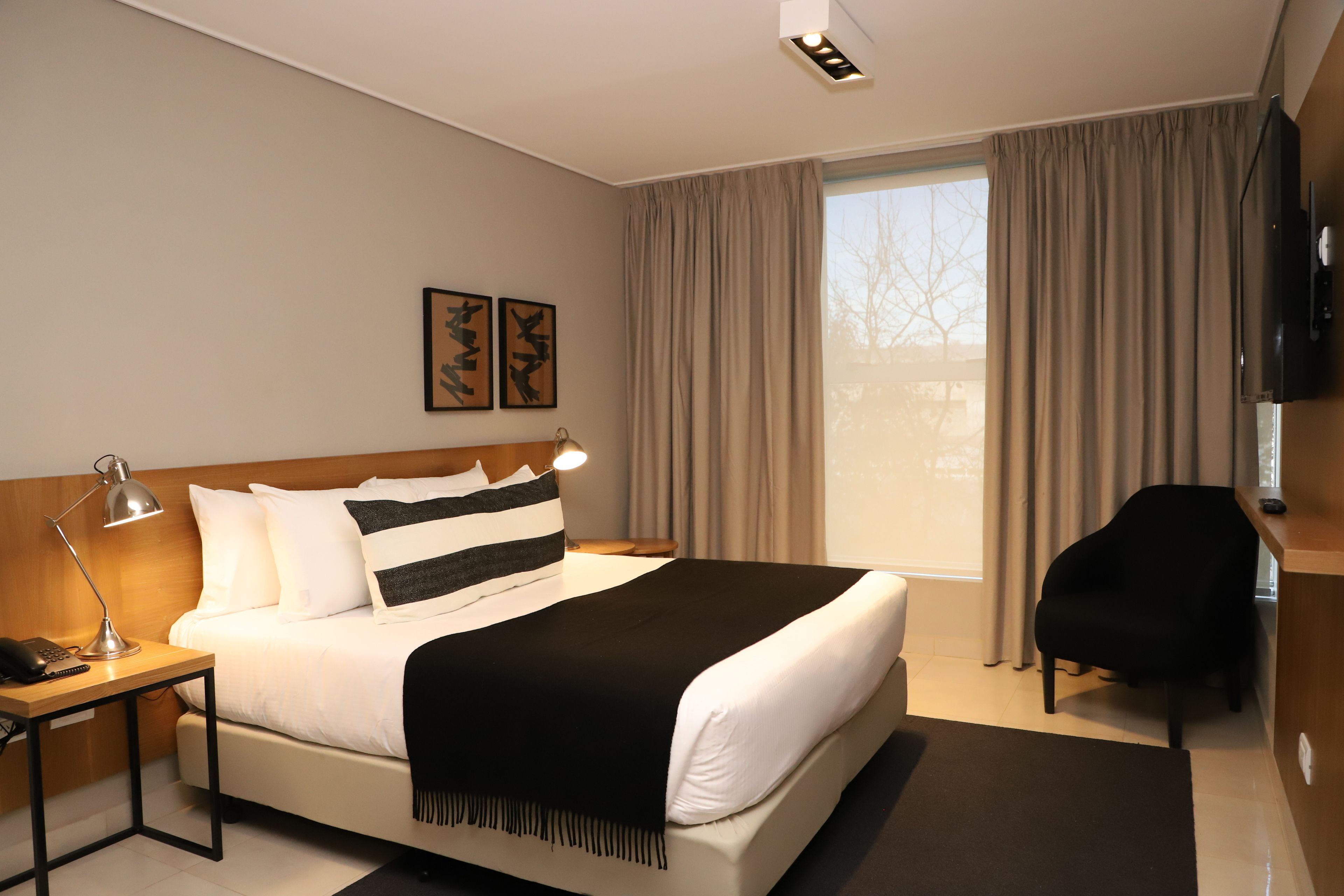 Una habitación de hotel moderna con una cama grande, un escritorio con lámpara, una silla y un televisor montado en la pared.