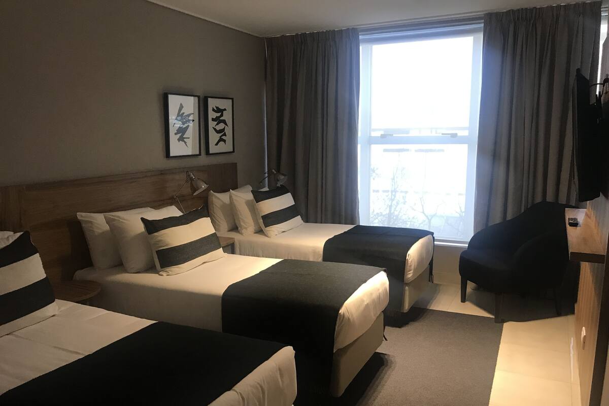 Ett hotellrum med två sängar, ett skrivbord, en stol och ett stort fönster med gardiner.